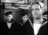 Four sailors in uniform stand together on a ship, with one man in the foreground looking serious while others watch attentively in the background. The black and white scene captures the intense atmosphere of a British naval war drama featured in “Glory at Sea (1952)” DVD, evoking the classic style of WWII minesweeper films.