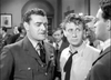 Two men, one dressed in a military uniform and the other in a shirt with a loose tie, stand close together in serious conversation at a crowded indoor gathering—reminiscent of scenes from the rare classic movie *Angels One Five (1952)*, now available on DVD. In the background, other people mingle and framed pictures adorn the walls.