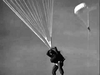A person in a parachute harness descends through the sky, holding parachute cords, echoing scenes from Operation Secret (1952) DVD, with another parachute visible against the clear sky.