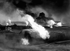 Large metal tanks and pipes are engulfed in flames, with thick smoke billowing into the sky—a chaotic scene of fire and destruction spreading throughout what appears to be an oil or industrial facility, reminiscent of the intense action and wartime drama depicted in the 1958 film "Hell Squad," now available on DVD.