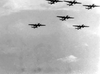 Black-and-white photo showing a group of military airplanes flying in formation across a cloudy sky, viewed from below—evoking the historical atmosphere of World War II as seen in classic films like Hell Squad (1958), now available on DVD.