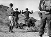 Four soldiers with raised hands surrender to an armed group in a rocky, desert landscape—a tense scene reminiscent of the 1958 war film "Hell Squad." One person lies on the ground while a soldier kneels beside them, capturing the dramatic urgency featured in this rare classic DVD. Two other figures observe the unfolding events, partially shown from behind.