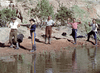 Five men stand with their hands raised beside a body of water in a desert landscape, surrounded by shrubs and rocky cliffs. Some wear cowboy hats and boots, bringing to mind a classic western scene reminiscent of rare films like *Southwest Passage (1954)* on DVD.