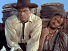 Two men in old-fashioned western clothing sit outdoors; one wears a wide-brimmed hat and looks serious, while the other looks exhausted, leaning back against wooden barrels beneath a blue sky. Capturing the gritty spirit of rare classic films, this scene evokes the timeless allure found in hard-to-find westerns like Southwest Passage (1954) DVD.