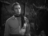 A man wearing a sheriff’s badge and neckerchief stands outdoors at night, gazing thoughtfully at a horse before him. Trees and a wooden structure can be seen in the background. This striking black and white image perfectly captures the classic western atmosphere of *Along the Great Divide (1951)*, making this DVD an ideal choice for fans of rare movies.