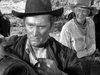 A serious man in a cowboy hat examines a saddle in the foreground, while an older man wearing a hat and bandana stands and smiles behind him. Captured outdoors with horses nearby and rendered in classic black and white, the scene feels straight out of “Along the Great Divide (1951) DVD”—a timeless moment that belongs in any classic western collection.