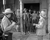 Four men in cowboy hats and western attire stand on a wooden sidewalk while two others, also wearing hats—one holding a rifle—face them. A horse is partly visible on the left. The scene, presented in striking black and white, captures the tense atmosphere of Badman’s Country (1958)—now available as a classic western DVD for collectors of rare movies on DVD.