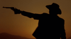 A cowboy in silhouette aims a revolver with his arm outstretched against an orange sunset sky, capturing the dramatic Western atmosphere featured in the “Dust (2001) DVD.”.
