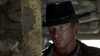 A man in a black cowboy hat and rugged clothing stares intently forward, set against a blurred backdrop of stone or wood that evokes the atmosphere of the Old West—an ideal scene for admirers of films like Dust (2001) DVD who are seeking to add an art-house western to their collection.
