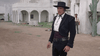 A man in black Western attire and a wide-brimmed hat stands in a dusty courtyard with white adobe buildings and cacti, evoking the atmosphere seen in 1960s western films like “Five for Revenge (1966)”, while another man observes from behind.