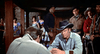 A tense scene in a Western saloon: two men sit at a table in the foreground, while others, both men and women, stand watching in the background. All wear period Western clothing, perfectly capturing the suspense and atmosphere reminiscent of Robber’s Roost (1955)—an unforgettable moment brought to life on DVD for classic western enthusiasts.