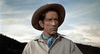 A man in a cowboy hat and blue neckerchief stands outdoors, looking serious—reminiscent of a character from Robber’s Roost (1955) DVD. The sky is partly cloudy, with hills stretching across the background.