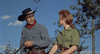 A man in a cowboy hat and red bandana rides a horse beside a woman in a green shirt, outdoors under a blue sky with trees behind them. Both gaze at each other, capturing the spirit of the classic western adventure seen in Robber’s Roost (1955) DVD.