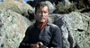A man in a dark shirt and red neckerchief stands outdoors among towering rocks, gun in hand and a serious expression on his face—evoking the intense atmosphere of “Robber’s Roost (1955) DVD” and making this moment an ideal addition to any classic western film collection.
