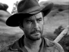 A bearded, mustached man in a wide-brimmed hat and rugged shirt stands outdoors in a classic western setting, gazing slightly to the side—evoking the timeless style of “Rocky Mountain (1950)” on DVD. The image is black and white.