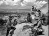 Six cowboys in hats and western attire gather on rocky terrain beneath a cloudy desert sky. Some are sitting, others standing, with one cowboy holding a rifle as saddles and gear lie scattered around them—capturing the rugged spirit of the Civil War western scene from Rocky Mountain (1950) on DVD.