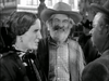Three older adults in Western attire converse outside. The woman on the left wears a dress and bonnet, the bearded man in the center smiles, and the man on the right wears glasses and a hat. The cheerful mood is reminiscent of classic western films like “Trail Street (1947) DVD.”.