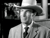 A man in a suit and cowboy hat stands indoors, looking slightly to the side with a neutral expression, channeling the timeless western style featured in Trail Street (1947), now available on DVD. The image is presented in black and white.