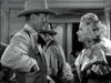 A man in a cowboy hat and a woman with flowers in her hair, dressed for the occasion, smile at each other in a lively saloon scene from the black-and-white classic western film “Trail Street (1947)”, available on DVD.