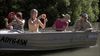 Four people sit in a small boat on a river, evoking the tense atmosphere of the film “Black Water (2007)” available on DVD. Two women are holding colorful rings, one woman rows at the front, and a man in a hat sits at the back, with dense bushes lining the water behind them.