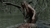 A disheveled person stands by a bloodied body near a riverbank, surrounded by dense trees and water, holding a sharp object—a chilling scene straight out of Black Water (2007), now available on DVD.