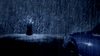 A person in a dark cape stands in heavy rain, facing a wall of bats illuminated by blue light. The surreal scene recalls the cover of the “Undead (2003) DVD,” an Australian horror-comedy, with a blue car parked in the foreground to the right.