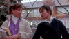 Two young women walk outdoors in autumn, wearing jackets and holding The Stepfather (1987) DVD as they chat. Behind them are a school building and trees with orange leaves.