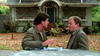 Two men converse before a boarded-up house with a blue door, checking papers on a car hood—one smokes as the other gestures. Fallen leaves cover the ground. A scene reminiscent of The Stepfather (1987) DVD, cult horror classic.