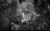 A black-and-white scene shows people gathered outdoors near a large circular stone—some standing, others kneeling or walking—surrounded by foliage and trees, creating a ritualistic, ceremonial atmosphere reminiscent of classic cult films. This intriguing image perfectly captures the chilling ambiance of rare hard-to-find movies such as “The Woman Eater (1958),” now available on DVD for British horror collectors.
