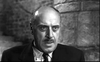 A serious-looking man with a mustache and receding hairline, dressed in a suit and tie and standing in front of a stone wall in this black-and-white image, evokes the enigmatic atmosphere of cult classic movies like “The Woman Eater (1958)”—a rare hidden gem now available on DVD.