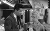 A man and a woman stand together at a carnival, illuminated by bright lights against a circus-themed backdrop. The woman gazes downward, seemingly deep in thought, while the man holds a drink and watches her. Captured in black and white, the image conjures the mysterious allure of cult classic cinema—much like The Woman Eater (1958) DVD, one of those rare hard-to-find movies prized by collectors.