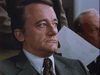A man in a suit and tie sits indoors, looking serious and slightly to the side, as if contemplating a crucial decision straight out of Virus (1999) on DVD—a suspenseful sci-fi horror thriller. Another person behind him holds a piece of paper, adding to the tension-filled atmosphere. The formal setting suggests a pivotal moment in an office or meeting environment, evocative of scenes from this classic film.
