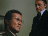 Two men are indoors; one in military uniform with glasses looks serious in the foreground, while another man in a suit stands behind him, partially in shadow. The tense atmosphere of this scene captures the chilling suspense of classic sci-fi horror thrillers like *Virus (1999)*, now available on DVD.