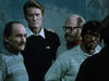Four men in sweaters and glasses stand indoors, one wearing a dark shirt with gold stripes on the shoulders. All look serious, with neutral or somber expressions, facing different directions against a muted blue-gray background—evoking the tense atmosphere of a sci-fi horror thriller like Virus (1999) on DVD, cherished by true collectors.
