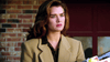 A woman with wavy brown hair wearing a tan blazer stands in front of a brick wall, looking slightly concerned or serious, as if she’s just stepped out of “Stalking Laura (1993) DVD”—one of those rare classic movies that keeps you guessing until the end.