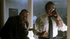 Two men in suits stand in a dimly lit office; one is speaking on a corded phone while the other leans in, both appearing serious and contemplative—a tense moment evocative of scenes from “Stalking Laura (1993)” DVD—while shadowy figures are visible through the window in the background.