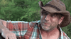 A man in a brown woven cowboy hat and plaid shirt smiles outdoors against a backdrop of green foliage—evoking the vibe of a character from “Clownado (2019)” on DVD, a cult horror-comedy splatter film featuring killer clowns.