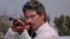 A man in a white shirt aims a shotgun, looking intently ahead—evoking the gritty energy of an ’80s sci-fi action thriller. Trees and a clear sky form the backdrop, perfectly capturing the dramatic atmosphere found in cult classics like The Hidden (1987) DVD.