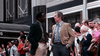 Two men in suits talk on a city sidewalk while a crowd of people, some looking on curiously, stand behind them near a modern building—perhaps discussing “The Winged Serpent (1982) DVD” or the latest rare movie releases as the city bustles around them.