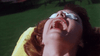 A person with reddish hair reclines outdoors, eyes shielded by white pads, mouth agape as if screaming or gasping, reminiscent of a scene from “The Winged Serpent (1982),” set against a lush green grassy backdrop.