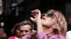 A woman in sunglasses and a purple top stands outdoors, tilting her head back and raising her hand to her mouth in a dramatic gesture reminiscent of a scene from “The Winged Serpent (1982)” DVD, with blurred people and buildings in the background.