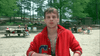 A young man in a red jacket stands on a sandy beach, looking directly at the camera. People are seated at picnic tables and on lawn chairs in the background, with trees and parked cars nearby—evoking the charged, cinematic atmosphere of classic thrillers like “The Sender (1982)” on DVD, where everyday moments brim with suspense.