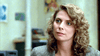 A woman with curly blonde hair wearing a gray blazer looks slightly to the side with a neutral expression, sitting indoors with blurred colorful artwork and a DVD of “The Sender (1982)” in the background.
