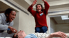 Two women laugh and shout while playfully using defibrillator paddles on a man lying on a hospital bed. One woman leaps excitedly, and the scene is chaotic yet humorous—evoking those unforgettable moments of comedy and pandemonium found in classic films like The Sender (1982) DVD.