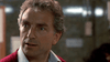 A man with curly gray hair, wearing a collared shirt and red sweater, looks surprised or concerned in an indoor setting with a blurred background—almost as if he’s just stumbled upon The Sender (1982) DVD or uncovered the chilling climax of a psychological horror film.