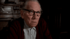 An elderly man with glasses and a maroon sweater sits indoors, gazing upward with a serious expression in a dimly lit room filled with wooden furniture—an atmosphere evocative of *The Tenant (1976) DVD* and the moody scenes often found in Roman Polanski’s films.