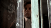 A man with short dark hair gazes out from behind a sheer lace curtain, his face partly hidden by the fabric and a blue window frame. Dressed in a collared shirt and wearing a serious expression, he evokes the haunting atmosphere of Roman Polanski’s The Tenant (1976) DVD.