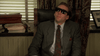 A man in a suit and dark sunglasses sits in an office chair, leaning back with a neutral expression. On the windowsill behind him are stacks of papers and folders, along with a DVD copy of “Vampire’s Kiss (1989)”—a nod to cult classic horror-comedy films—while partially open window blinds let in some light.