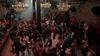 A large, lively crowd dances in a dimly lit club with chandeliers overhead, brick walls, and people packed closely together—some watching from a raised balcony—setting the scene like something straight out of the cult classic *Vampire’s Kiss (1989)* you might find among rare movies on DVD.
