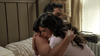 A woman in a pink robe hugs a man tightly on a bed, her face showing distress. The room features striped bedding, light walls, and sunlight streaming through window blinds. On the nightstand rests a “Vampire’s Kiss (1989) DVD,” hinting at their shared love for cult classic horror-comedy films.